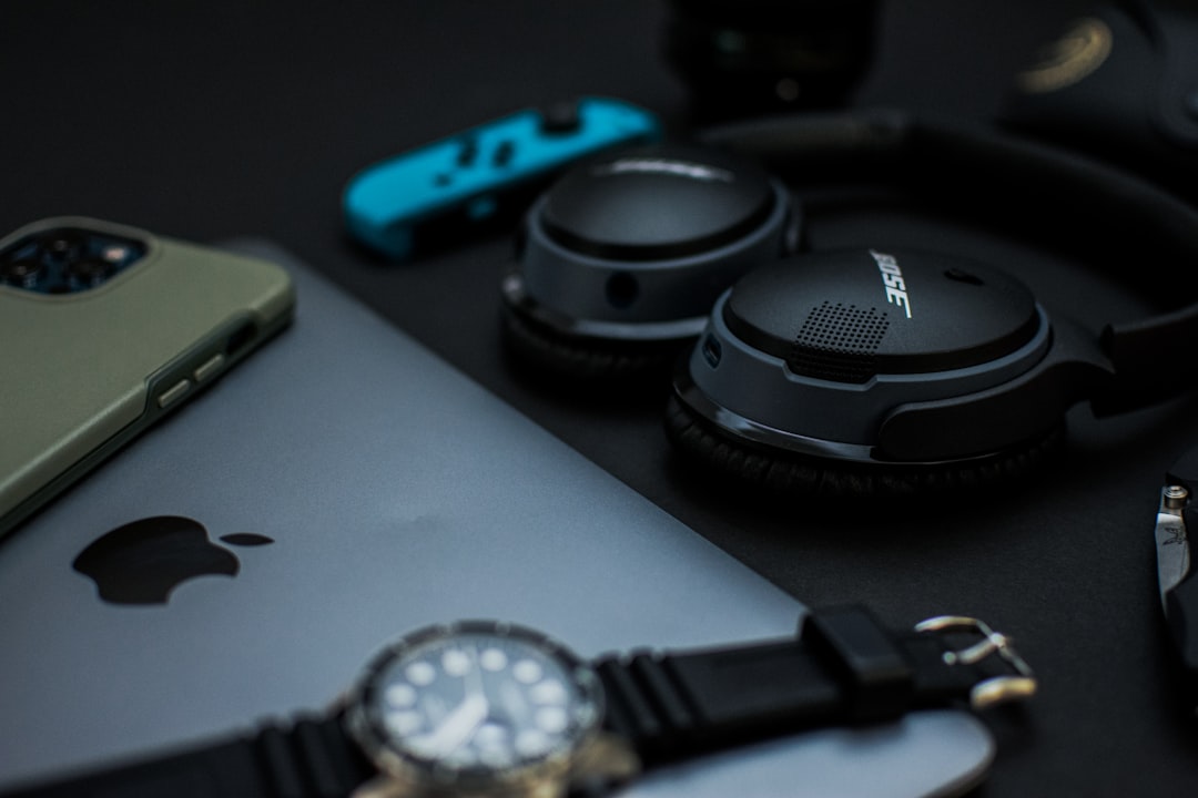 Flat lay headphones, watch, sunglasses, knife, laptop, phone, joycon, lens, and camera on black backdrop
